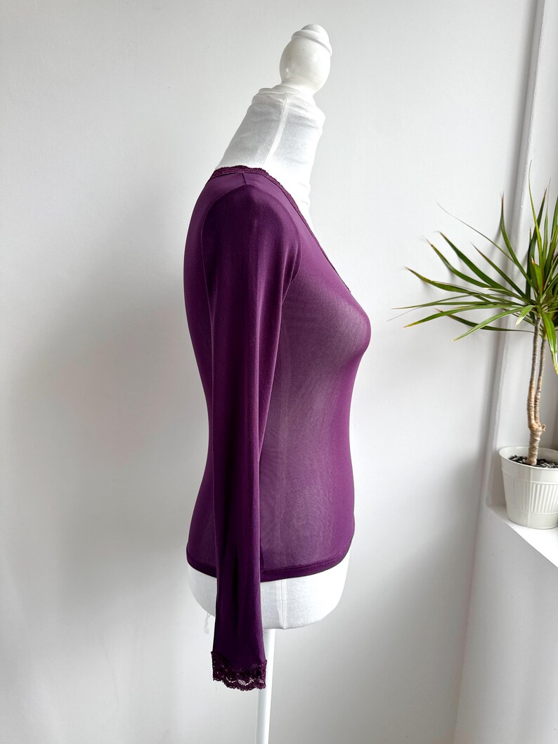 May include: A long-sleeved, deep purple top with a V-neck and delicate lace trim at the cuffs and neckline. The sheer fabric drapes over a white mannequin, showcasing the garment's form and design. The top is a stylish addition to any wardrobe.