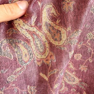 May include: Close-up of a woven textile with a paisley pattern. The fabric is primarily a deep rose color with accents of gold, tan, and black. The intricate design is visible in the folds of the material.