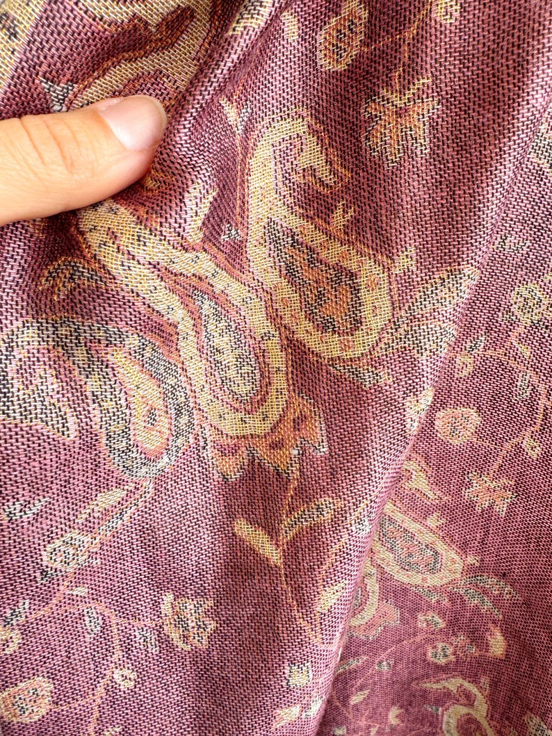 May include: Close-up of a woven textile with a paisley pattern. The fabric is primarily a deep rose color with accents of gold, tan, and black. The intricate design is visible in the folds of the material.
