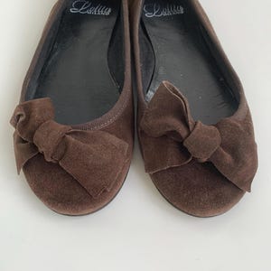 May include: A pair of brown suede ballet flats with bow accents. The shoes have a rounded toe and a flat sole. The brand name "Lolitus" is printed in cursive on the insole. The shoes are displayed on a white surface.