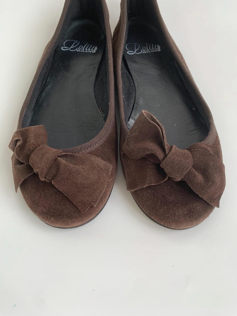 May include: A pair of brown suede ballet flats with bow accents. The shoes have a rounded toe and a flat sole. The brand name "Lolitus" is printed in cursive on the insole. The shoes are displayed on a white surface.