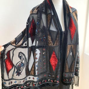 Op de afbeelding: Een vintage zwarte sjaal met een geometrisch patroon van ruiten en vierkanten in tinten grijs, bruin en rood. De sjaal heeft een fluwelen rand en franjes langs de onderkant. De sjaal wordt op een mannequin getoond.