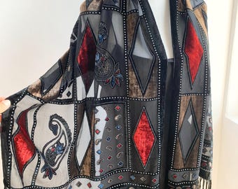 May include: A vintage black scarf with a geometric pattern of diamonds and squares in shades of gray, brown, and red. The scarf has a velvet trim and fringe along the bottom edge. The scarf is displayed on a mannequin.