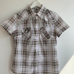 Puede incluir: Una camisa blanca de manga corta con botones y un estampado de cuadros verde, blanco y morado. La camisa tiene dos bolsillos en el pecho con cierre de botón.