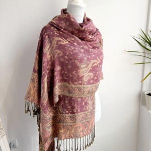 May include: A burgundy paisley scarf with a cream and gold border and fringe. The scarf is draped over a white mannequin. The paisley pattern is a repeating design of curved, teardrop-shaped motifs. The scarf is made of a soft, flowing fabric.