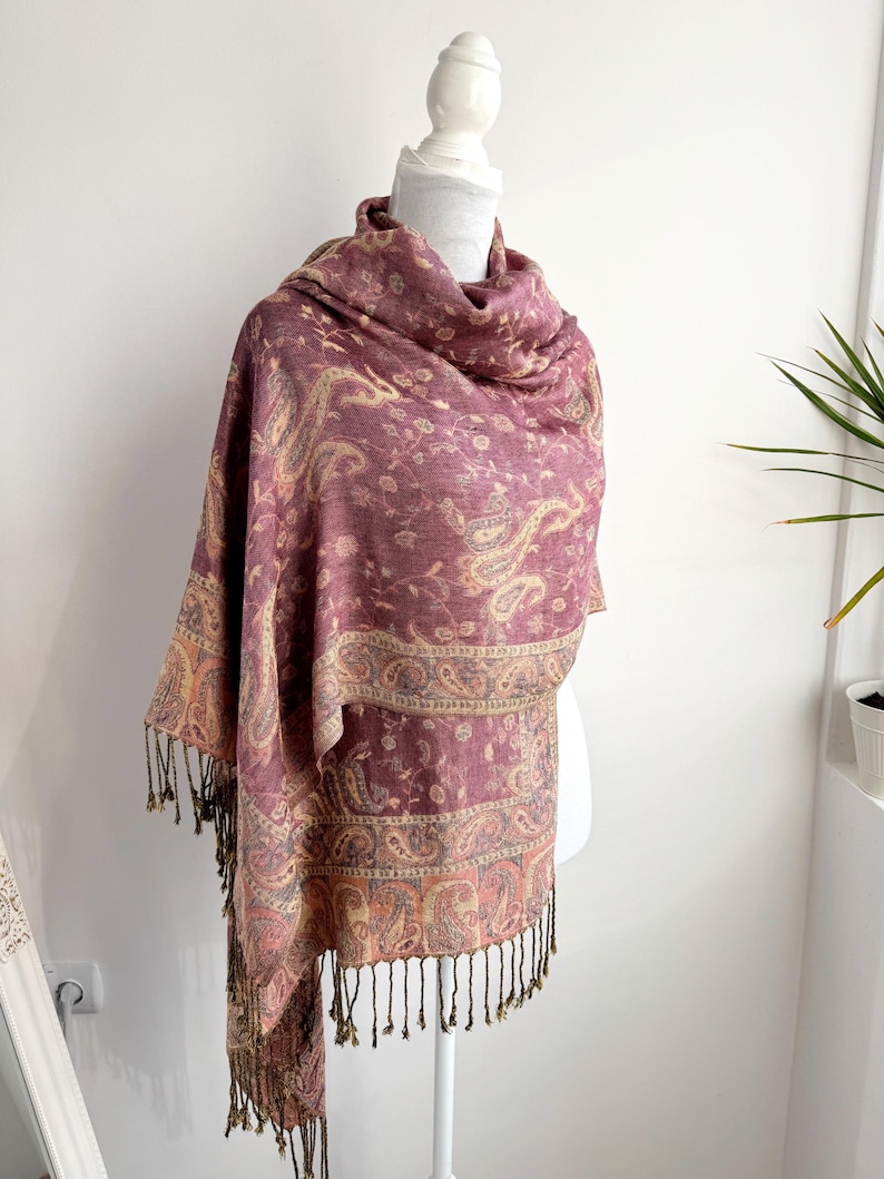 May include: A burgundy paisley scarf with a cream and gold border and fringe. The scarf is draped over a white mannequin. The paisley pattern is a repeating design of curved, teardrop-shaped motifs. The scarf is made of a soft, flowing fabric.