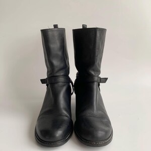 May include: A pair of black leather mid-calf boots featuring a rounded toe, a stacked heel, and a thin adjustable strap with a buckle detail wrapped around the ankle area. The boots have a smooth finish and a pull tab at the top of the shaft.