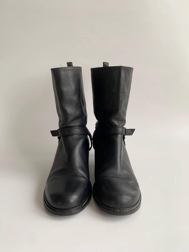 May include: A pair of black leather mid-calf boots featuring a rounded toe, a stacked heel, and a thin adjustable strap with a buckle detail wrapped around the ankle area. The boots have a smooth finish and a pull tab at the top of the shaft.