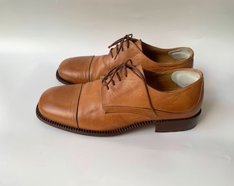 May include: A pair of brown leather lace-up shoes with a square toe. The shoes have a brown leather sole and a brown leather heel.