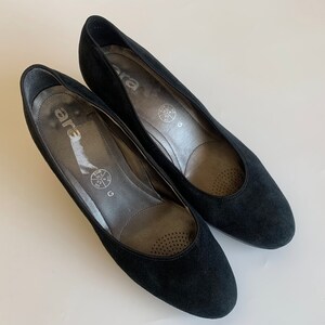 May include: A pair of black suede pumps with a low heel. The shoes have a round toe and a simple design. The brand name "ara" is printed on the insole.