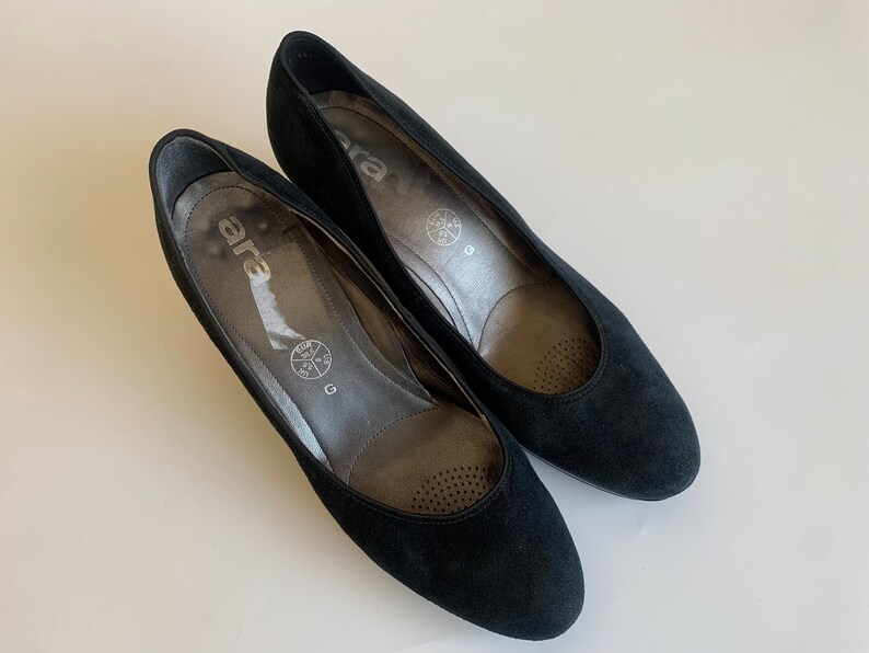 May include: A pair of black suede pumps with a low heel. The shoes have a round toe and a simple design. The brand name "ara" is printed on the insole.