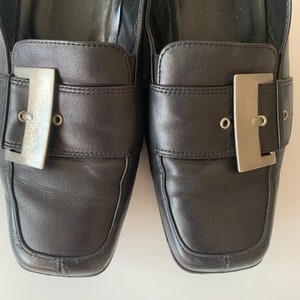 K&ouml;nnte beinhalten: Ein Paar schwarze Leder-Slipper mit silbernen Schnallen. Die Slipper haben eine quadratische Spitze und einen niedrigen Absatz.