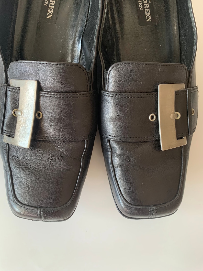K&ouml;nnte beinhalten: Ein Paar schwarze Leder-Slipper mit silbernen Schnallen. Die Slipper haben eine quadratische Spitze und einen niedrigen Absatz.