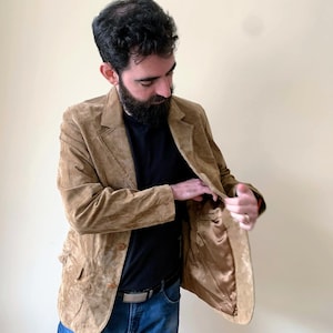 May include: A brown suede blazer with a three-button closure. The blazer has a brown satin lining and a pocket on the inside.