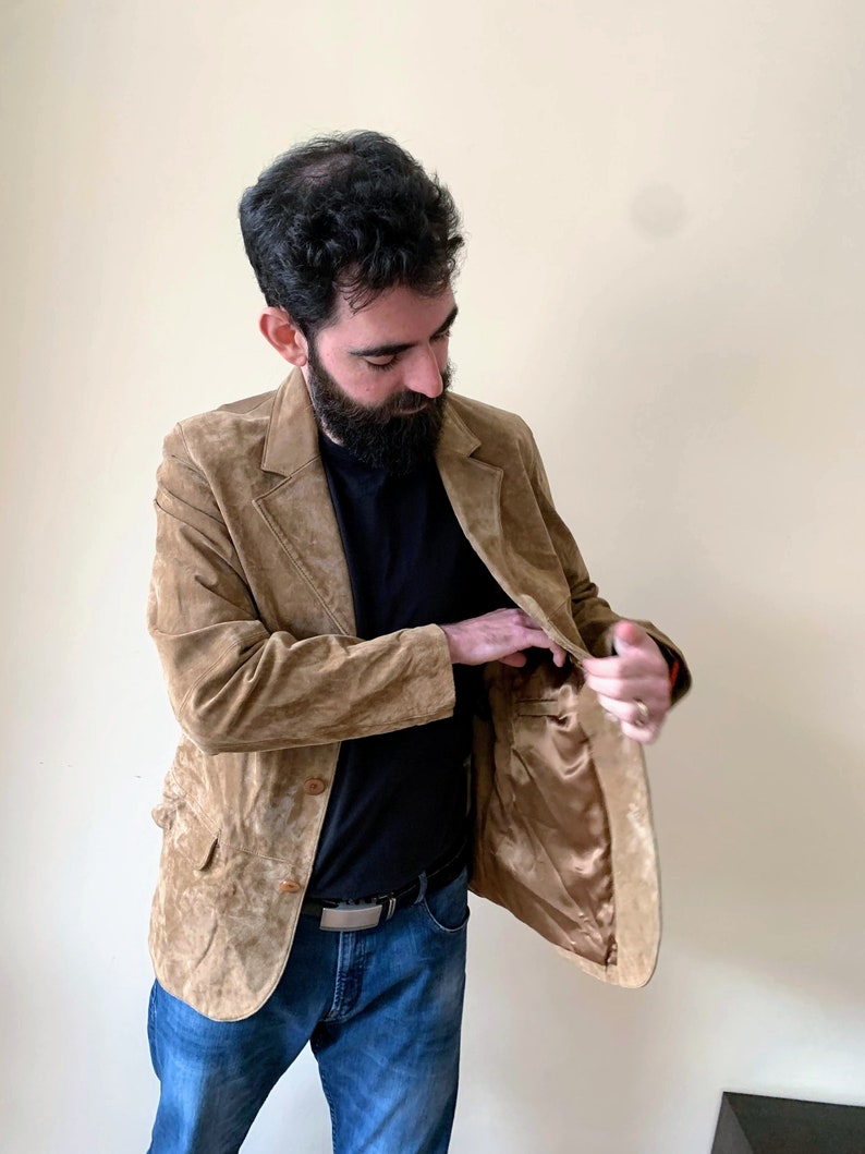 May include: A brown suede blazer with a three-button closure. The blazer has a brown satin lining and a pocket on the inside.