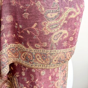 May include: A close-up of a burgundy shawl with a detailed paisley pattern in beige, tan, and gray. The shawl has a decorative border with intricate designs. The fabric appears soft and woven.