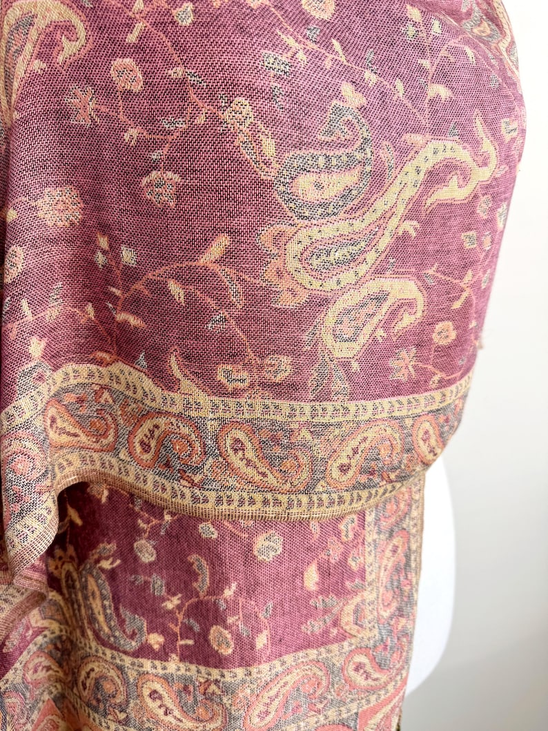 May include: A close-up of a burgundy shawl with a detailed paisley pattern in beige, tan, and gray. The shawl has a decorative border with intricate designs. The fabric appears soft and woven.