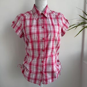 Può includere: Una camicia a quadri rossi e bianchi a maniche corte, con chiusura a bottoni sul davanti e due piccole tasche sul petto. La camicia ha un colletto e lacci laterali regolabili. La camicia è esposta su un manichino bianco.