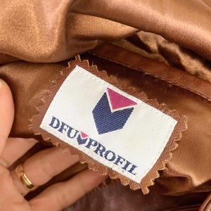 May include: Close-up of a brown leather garment with a satin lining. A label with the text "DFU PROFIL" and a geometric logo is sewn into the garment. The garment appears to be vintage.