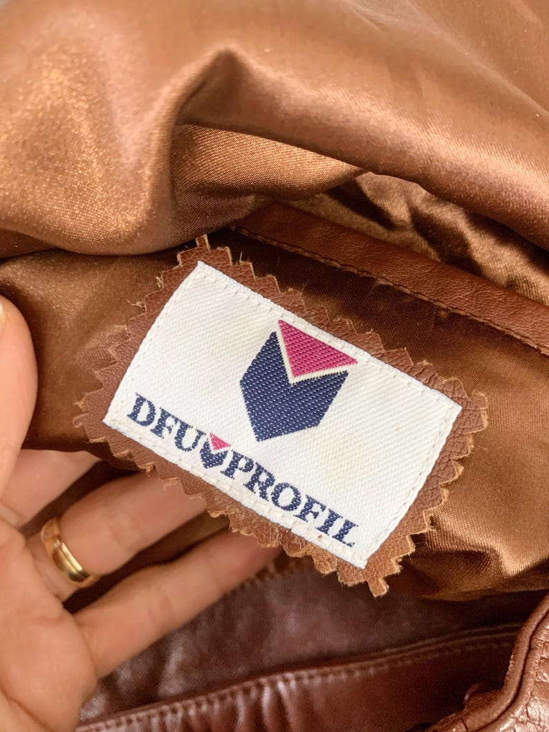 May include: Close-up of a brown leather garment with a satin lining. A label with the text "DFU PROFIL" and a geometric logo is sewn into the garment. The garment appears to be vintage.