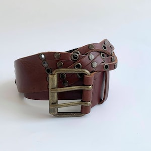 以下が含まれることがあります： 編み込みのデザインと金属製のスタッズが付いた茶色の革ベルト。ベルトには、ダブルプローブクロージャー付きの真鍮製のバックルが付いています。