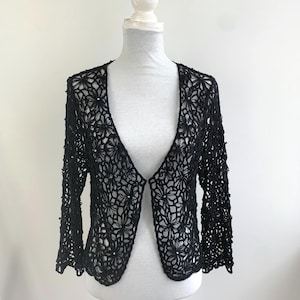 May include: Black open-front crochet cardigan with long sleeves. The floral pattern is visible throughout the garment. The cardigan is displayed on a white mannequin against a white background. The sleeves end just below the elbow.