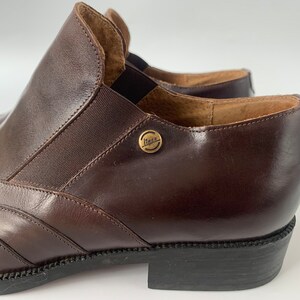 K&ouml;nnte beinhalten: Ein Paar braune Lederstiefeletten mit einem goldenen Metallschild mit der Aufschrift "Lita" an der Seite. Die Stiefeletten haben eine spitze Zehenpartie und einen niedrigen Absatz.