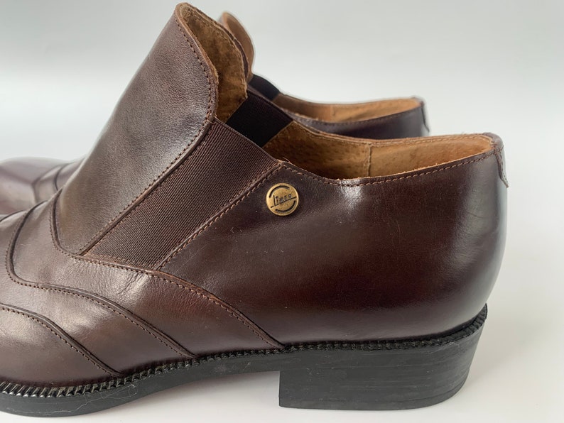 K&ouml;nnte beinhalten: Ein Paar braune Lederstiefeletten mit einem goldenen Metallschild mit der Aufschrift "Lita" an der Seite. Die Stiefeletten haben eine spitze Zehenpartie und einen niedrigen Absatz.