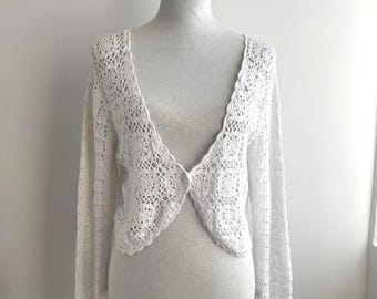 May include: A white, open-front crochet cardigan with long sleeves. The delicate, lacy pattern covers the entire garment, which is displayed on a mannequin. The cardigan has a single button closure at the waist.