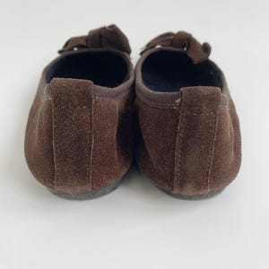 May include: A pair of brown suede ballet flats. Each shoe has a small bow on the top and a rounded toe. The shoes are set against a white background.
