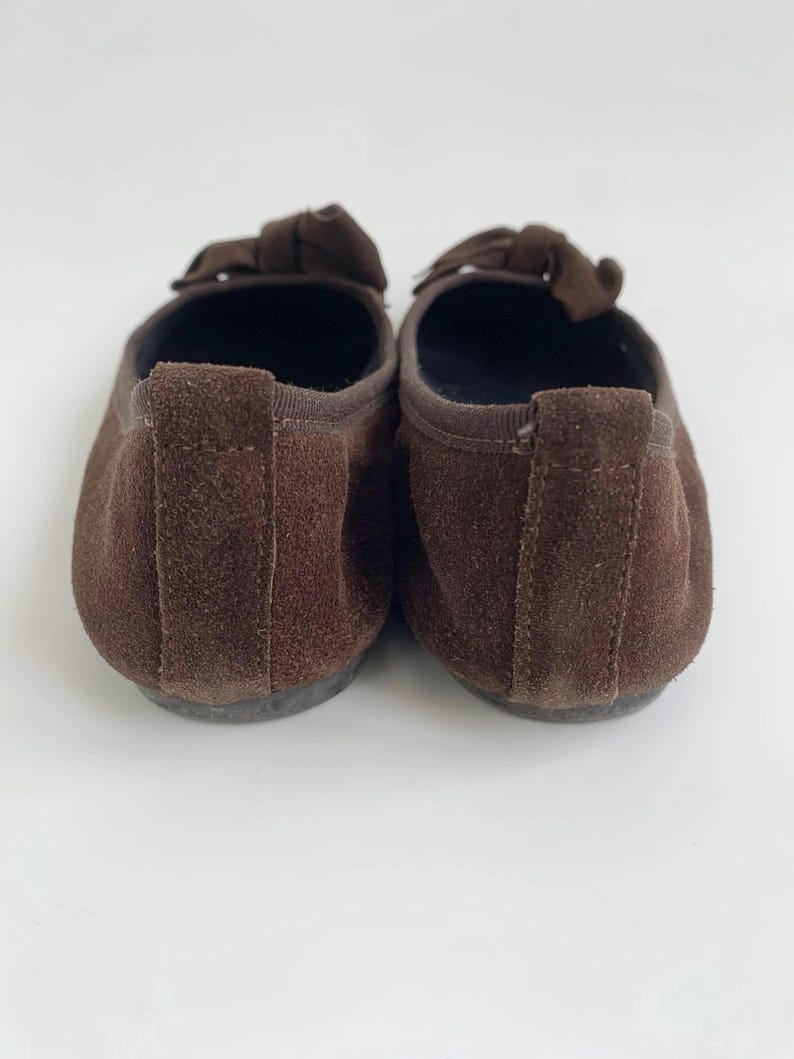 May include: A pair of brown suede ballet flats. Each shoe has a small bow on the top and a rounded toe. The shoes are set against a white background.