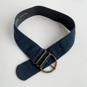 May include: A navy blue belt with a textured surface and a brass-coloured buckle. The belt is wide and has a simple, classic design. The text "FABRIQUE EN FRANCE" is printed on the inside of the belt.