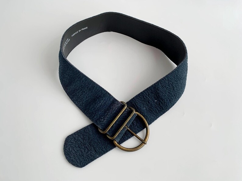 May include: A navy blue belt with a textured surface and a brass-coloured buckle. The belt is wide and has a simple, classic design. The text "FABRIQUE EN FRANCE" is printed on the inside of the belt.