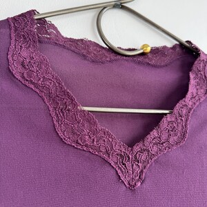 May include: A close-up of a purple top with a V-neckline. The neckline is trimmed with a delicate, dark purple lace. The top is hanging on a silver metal hanger with gold-colored accents.