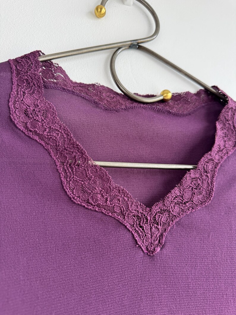 May include: A close-up of a purple top with a V-neckline. The neckline is trimmed with a delicate, dark purple lace. The top is hanging on a silver metal hanger with gold-colored accents.