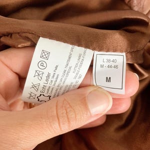 May include: Close-up of a brown leather garment's interior, revealing a care label with washing instructions and size information. The label indicates the garment is made of genuine leather and polyester. The size is indicated as M-44-46 and L 38-40.