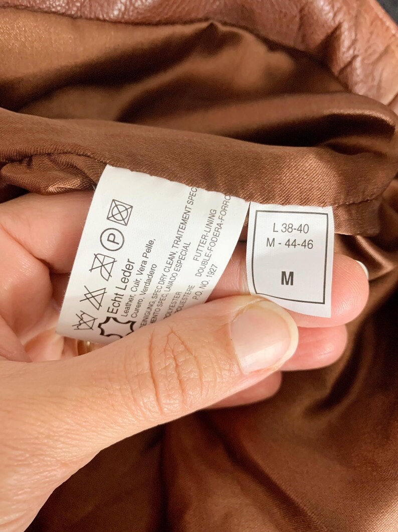 May include: Close-up of a brown leather garment's interior, revealing a care label with washing instructions and size information. The label indicates the garment is made of genuine leather and polyester. The size is indicated as M-44-46 and L 38-40.