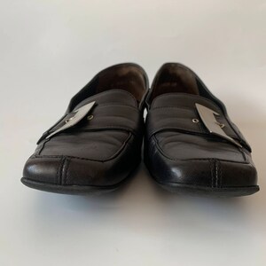 K&ouml;nnte beinhalten: Ein Paar schwarze Leder-Slipper mit silbernen Schnallen. Die Slipper haben eine quadratische Spitze und einen niedrigen Absatz.