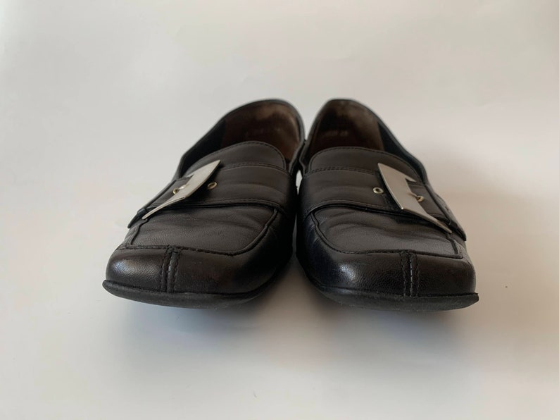 K&ouml;nnte beinhalten: Ein Paar schwarze Leder-Slipper mit silbernen Schnallen. Die Slipper haben eine quadratische Spitze und einen niedrigen Absatz.