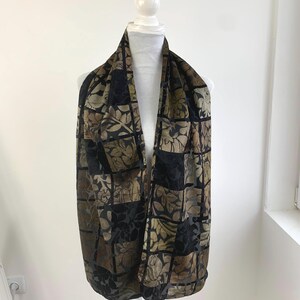 May include: A patterned scarf in black, brown, and olive green, draped over a white mannequin. The scarf features a repeating square design with floral motifs. The fabric appears soft and flowing, suitable for various styles. The scarf is a versatile accessory.