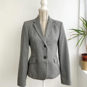 Könnte beinhalten: Ein grauer Blazer mit Zwei-Knopf-Verschluss, fallendem Revers und zwei Vordertaschen. Die Jacke wird auf einer Schaufensterpuppe vor weißem Hintergrund präsentiert. Der Blazer hat lange Ärmel und eine taillierte Passform.