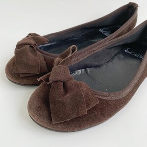 May include: A pair of brown suede ballet flats with large bow accents. The shoes have a rounded toe and a thin, flexible sole. The interior is black, and the shoes are set against a white background.