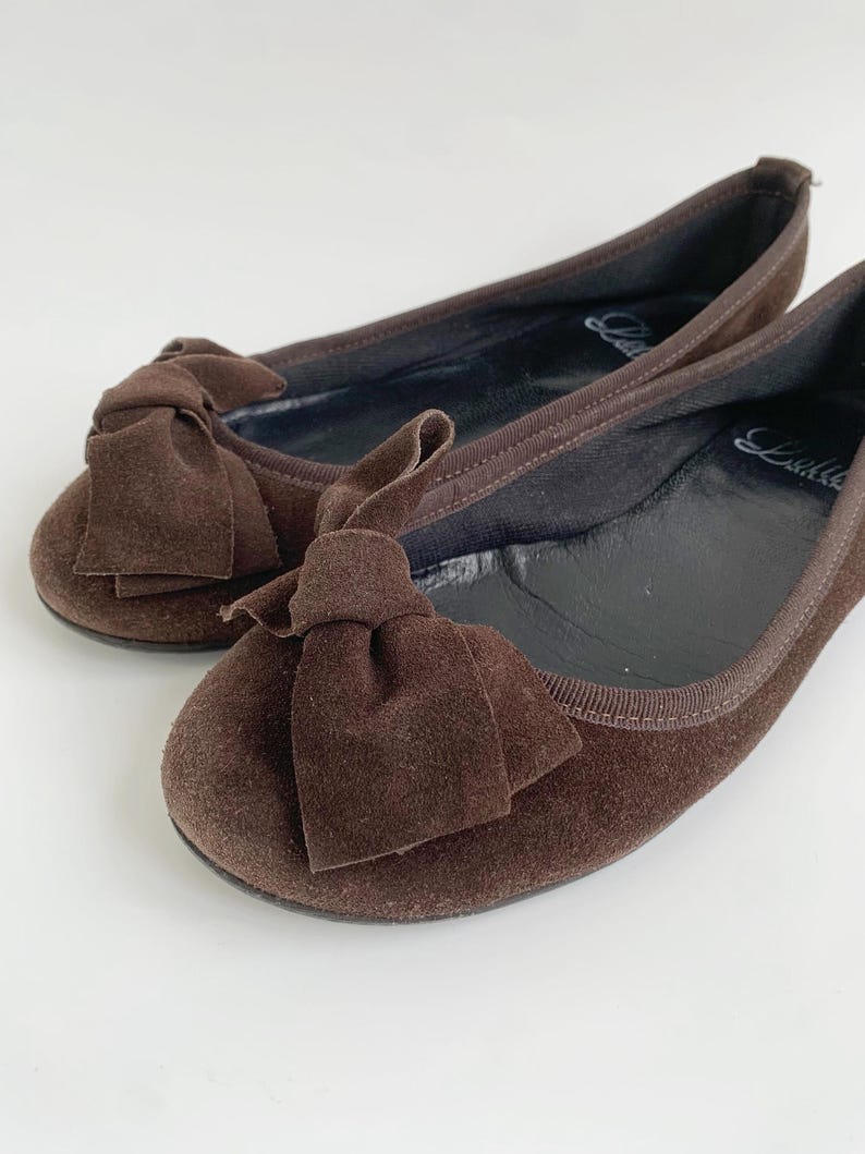 May include: A pair of brown suede ballet flats with large bow accents. The shoes have a rounded toe and a thin, flexible sole. The interior is black, and the shoes are set against a white background.