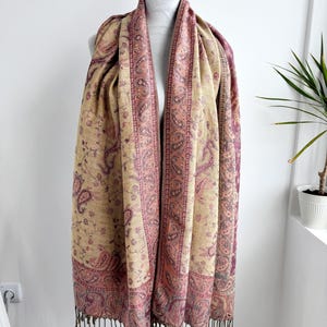 May include: A beige and burgundy paisley patterned scarf with a fringed edge. The scarf is draped over a white mannequin. The paisley design is in shades of burgundy and pink, with a beige background.
