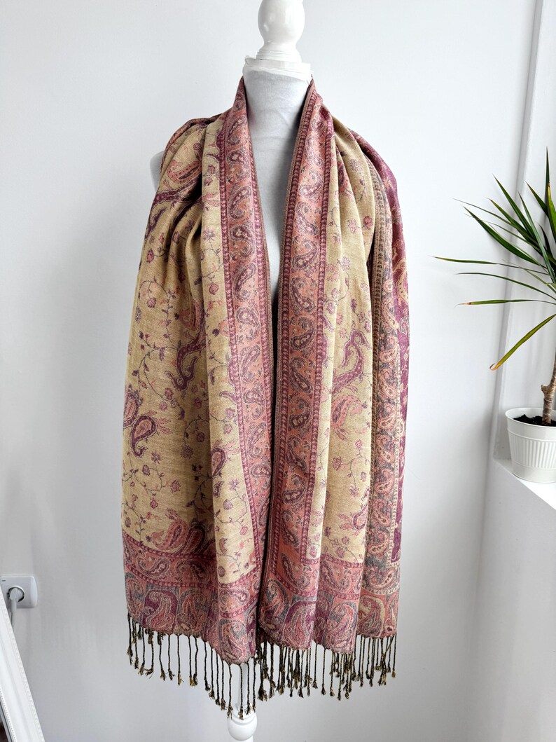 May include: A beige and burgundy paisley patterned scarf with a fringed edge. The scarf is draped over a white mannequin. The paisley design is in shades of burgundy and pink, with a beige background.