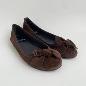 May include: A pair of brown suede ballet flats with a bow detail on the toe. The shoes have a rounded toe and a flat sole. The brand name "Callus" is visible on the insole. The background is plain white.