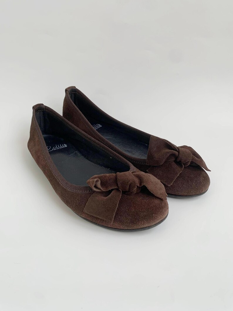 May include: A pair of brown suede ballet flats with a bow detail on the toe. The shoes have a rounded toe and a flat sole. The brand name "Callus" is visible on the insole. The background is plain white.