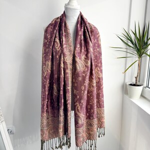 May include: A plum-colored scarf with a paisley pattern and fringe. The scarf is draped over a white mannequin. The paisley design includes beige and gold accents. The fringe is a dark olive green color.