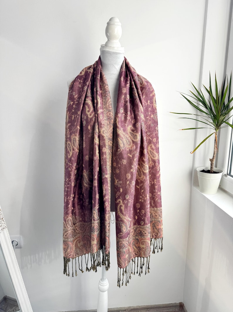 May include: A plum-colored scarf with a paisley pattern and fringe. The scarf is draped over a white mannequin. The paisley design includes beige and gold accents. The fringe is a dark olive green color.