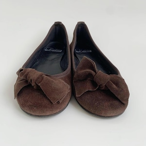 May include: A pair of brown suede ballet flats with large bow accents. The shoes have a rounded toe and a flat sole. The brand name "Lalita" is visible on the insole. The shoes are set against a white background.